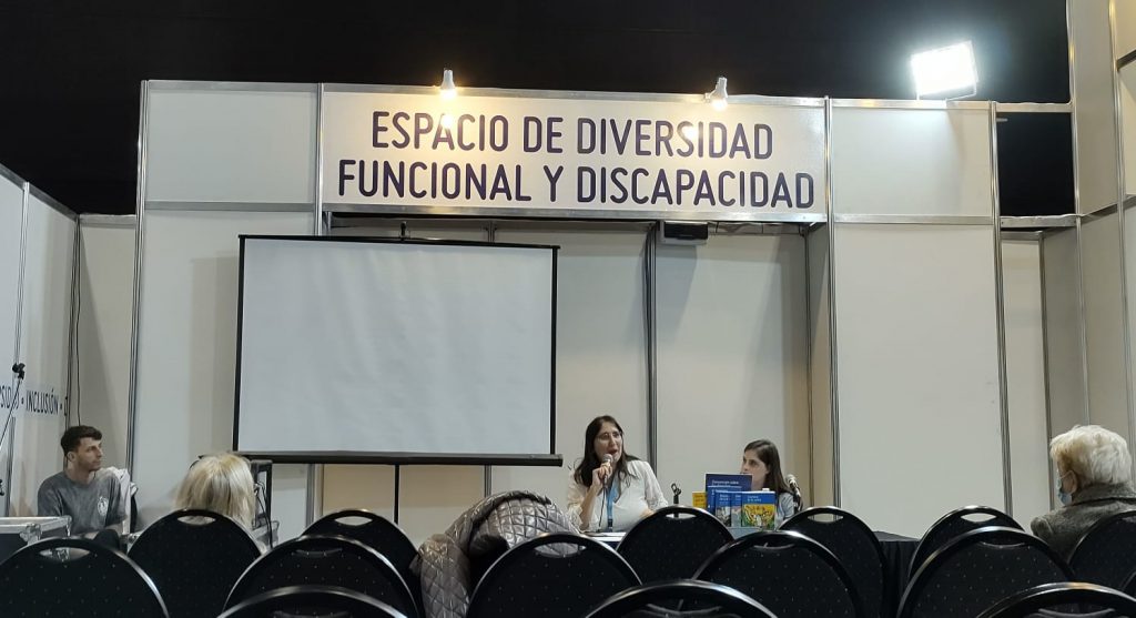 Conferencia. Se ve a los expositores de Fundación Visibilia. Dos mujeres, una de mediana edad hablando por micrófocno y a su lado una mujer joven con discapacidad