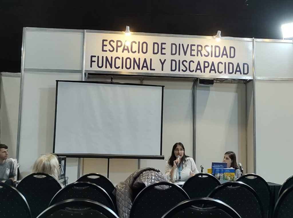 Conferencia. Se ve a los expositores de Fundación Visibilia. Dos mujeres, una de mediana edad hablando por micrófocno y a su lado una mujer joven con discapacidad