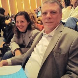 Dos personas , un varon y una joven, sentados en un auditorio, de costado, sonriendo. Ambos vestidos formalmente